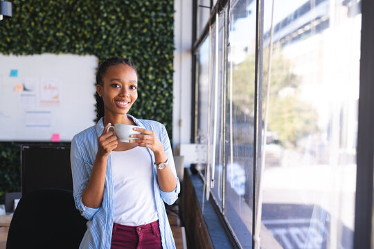 Happy african american casual businesswoman drinking coffee by window in office, copy space