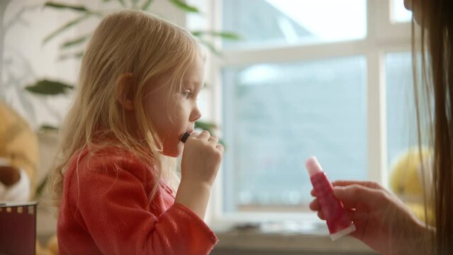 Cute toddler girl shows process of applying lipbalm on her lips, side view, beauty care, girl's growing. Family weekend, female beauty routine, sunny nursery room background. High quality 4k footage