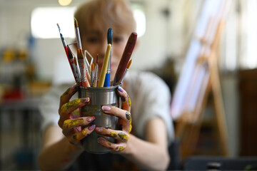 Image of male artist in apron holding metal bucket with different brushes of the artist. Art, education, creativity and people © Prathankarnpap