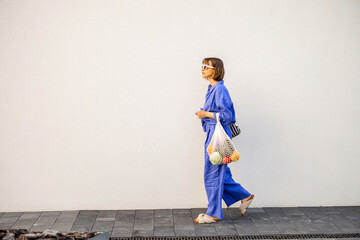 Woman in blue pajamas walks with mesh bag full of fresh fruits on white wall background outdoors....