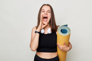 Healthy lifestyle choices. Sporty and active lifestyle. Excited woman holding rolled fitness mat dressed in activewear isolated over gray background screaming making announcement keeps hand near mouth