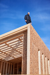 Male developer building wooden frame house. Bald man standing on construction site, inspecting quality of work on sunny day with blue sky on background.