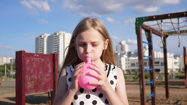child girl inflates a pink air balloon, summer portrait in city park