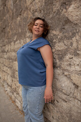 A beautiful girl is wearing a blue t-shirt and jeans, the overweight woman posing against stone biege wall. The fashion outfit for plus-size woman for the city, minimalizm urban style clothes