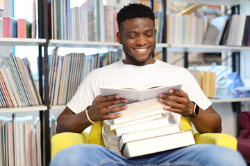 Smiling black young guy holding a stack of books in the library.