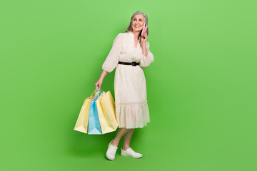 Full length photo of adorable cute lady wear beige dress talking gadget holding shoppers looking empty space isolated green color background
