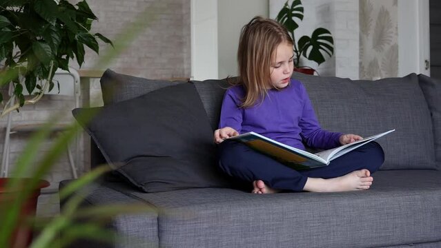 A cute little blonde girl is reading a large colorful book sitting on the sofa in the living room. Self-education of younger schoolchildren, reading literature at home, interesting leisure