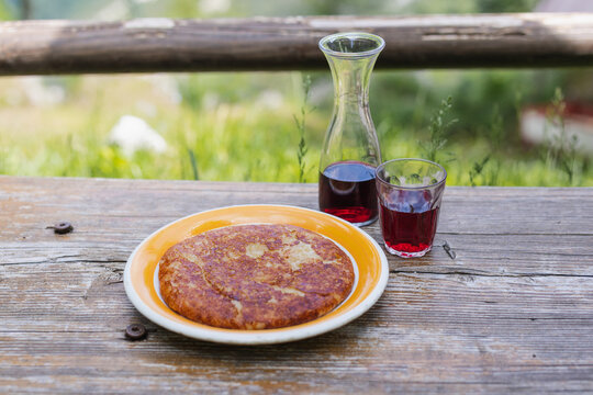 Un piatto di Frico, il tradizionale piatto del Friuli Venezia Giulia sfondo montagna. Una caraffa e un bicchiere di vino rosso disposti su un tavolo di legno grezzo.