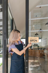 Young asian businesswoman small business owner standing at door entrance signboard, A cheerful entrepreneur young waitress in a blue apron near the glass door