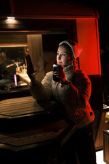 Model blonde with tails with cup of coffee near coffee shop with red and yellow light looking in smartphone. Girl weared white dress, jacket and hat posing near cafe in winter night