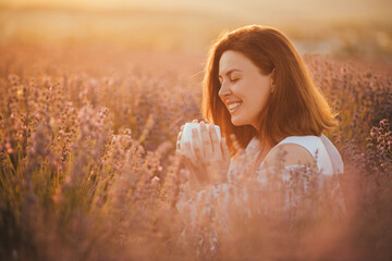 Young happy woman drinking herbal tea, sitting in a beautiful lavender field at sunset. - obrazy, fototapety, plakaty