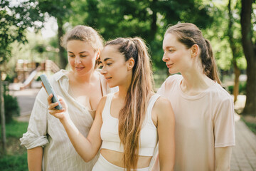 Diverse millennial women spending time together in a park, using smartphone.