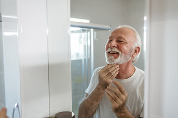 Men's personal care. Bearded senior man puts cream on his face in the bathroom.