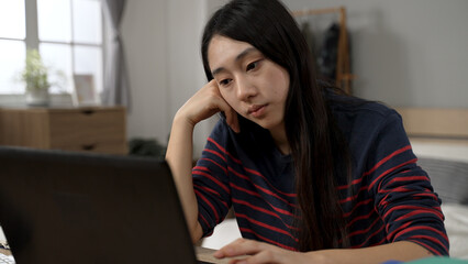 stressed Asian female adult student is propping head and looking at the computer screen while learning a difficult subject from home at desk.