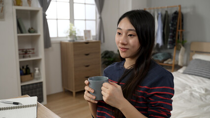 attractive Asian college girl learning from home is smiling into distance while enjoying tea and the tranquil morning at desk in her modern bedroom with daylight