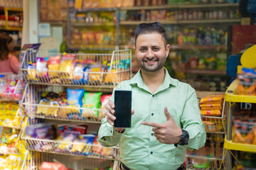 Young indian man showing smartphone screen at grocery shop.