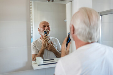 Bearded senior man in white shirt trimming his beard with a trimmer at home.