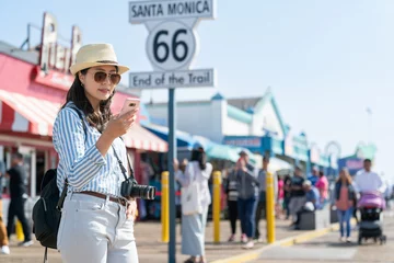 Fotobehang cheerful asian Chinese female tourist searching for best seafood restaurant on phone near route 66 end of the trail sign during spring holiday at santa monica pier in the us © PR Image Factory
