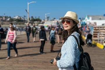 beautiful smiling asian korean girl wearing sunglasses and backpack turning to look at distance at santa monica pier with tourists coming and going at background