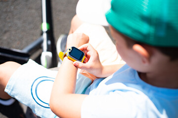 A little 7 year old boy in bright summer clothes looks at his yellow smart watch on his hand - 628123959