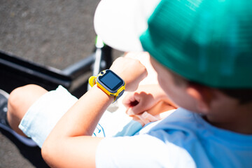 A little 7 year old boy in bright summer clothes looks at his yellow smart watch on his hand - 628123964