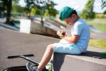 A little 7 year old boy in bright summer clothes looks at his yellow smart watch on his hand - 628123977