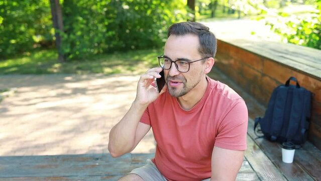 A man with glasses uses a phone, talking on a smartphone outdoors in a park on a summer day.