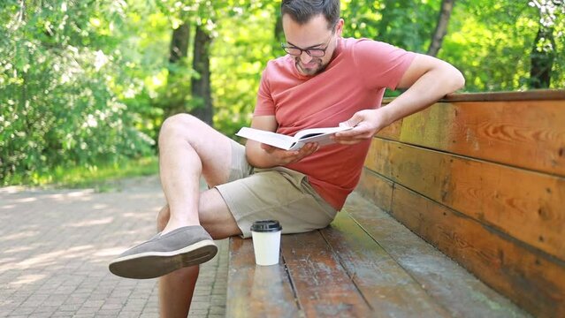 A man with glasses reads a book and drinks coffee while sitting on a park bench on a summer day.