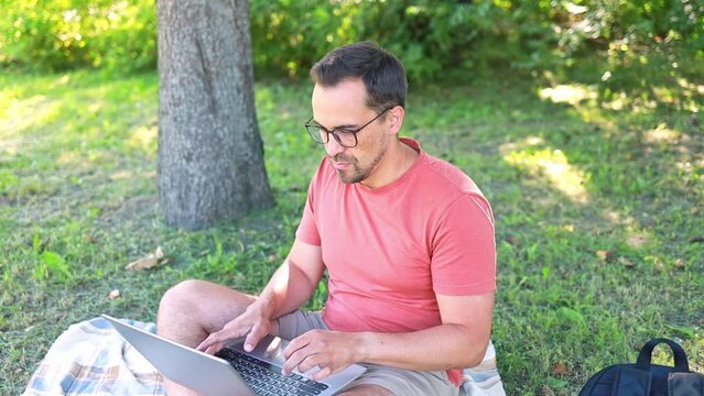 Remote work in the park. A man uses a laptop, typing text on the keyboard while sitting on a blanket on the grass in the park.
