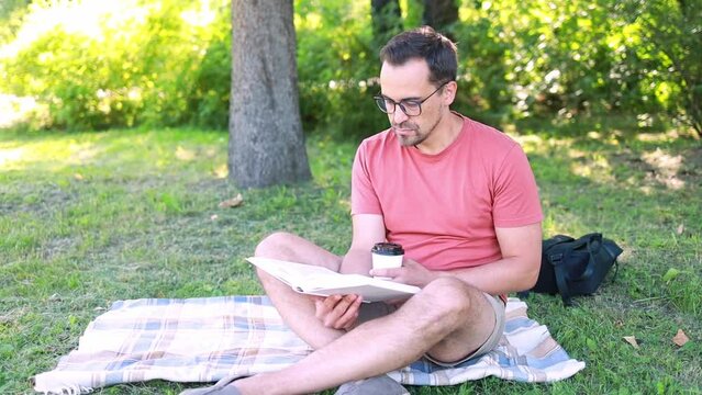 Camping. A man reads a book and drinks coffee while sitting on a blanket on the grass in a park on a summer day.