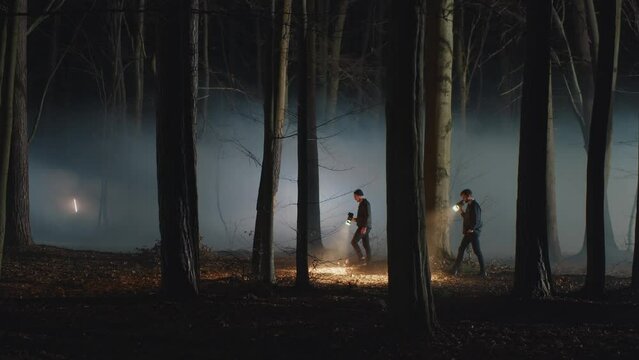 Two men walking in the night foggy forest with flashlights. Detectives searching clues or body among the trees. Crime scene, TV or movie experience, police work