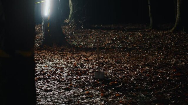 Shovel stuck in ground covered with wet fallen leaves, nighttime autumn forest, crime scene restricted by yellow police tape. Detective investigation process, police work, forensic experts