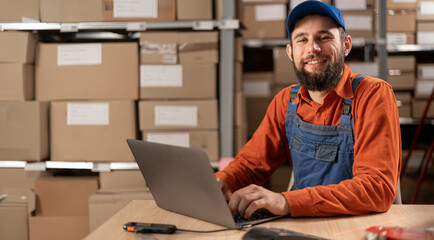 Portrait of a happy young male worker working on laptop in warehouse, Preparing parcels for delivery.