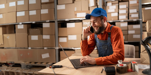 Warehouse worker doing inventory using laptop calling customers on mobile phone making a deal