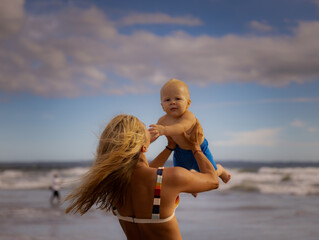 Mother enjoying summer vacations, holding, playing and lifting her infant baby boy son high in the air on sandy beach. Family travel and vacations concept. Seminyak beach, Bali