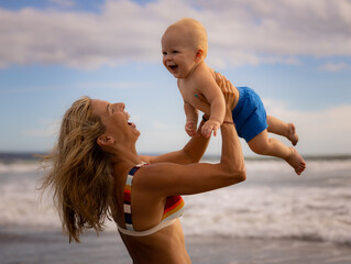 Mother holding and lifting her infant baby boy high in the air on beach. Happy family. Mum and son laughing and smiling. Blue sky with white clouds. Summer vacation. Seminyak beach, Bali