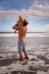 Father enjoying summer vacations, holding, kissing and lifting his infant baby boy son high in the air on sandy beach. Family travel and vacations concept. Seminyak beach, Bali