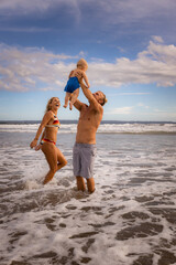 Young family spending summer vacation in Asia. Father holding, lifting his infant baby boy son high in the air. Happy mother smiling and laughing. Family beach concept. Seminyak beach, Bali