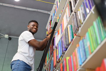 A black man on the stairs chooses a book from a bookshelf in the library. - Powered by Adobe