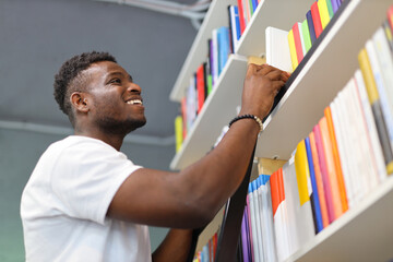 Black man reaching for a book on a bookshelf while wearing a white t-shirt and a black watch. The background is a gray wall and the bookshelf has multiple rows of colorful books.