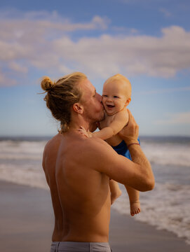 Father holding, kissing his infant baby boy son high in the air on sandy beach. Family travel and summer vacations concept. Family outdoor activities. Seminyak beach, Bali