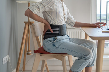 Man freelancer works types on computer keyboard wearing back support belt corset on lower back during exacerbation to treatment of hernia. Back pain health problems as consequences of sedentary work.  - obrazy, fototapety, plakaty