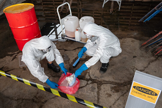 A team of two chemists, wearing PPE suits and gas masks, recover a deadly chemical spill on the factory warehouse floor. Correct disposal of chemical spills in industrial plants.