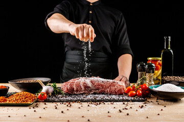 Chef applying grained salt on raw piece of steak. Cooker preparing meat on professional kitchen table with various vegetables 