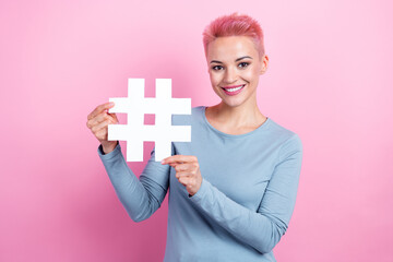 Portrait of pleasant lovely adorable woman with dyed hairdo wear stylish clothes hold hashtag icon isolated on pink color background