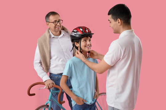 Young Man Putting Bike Helmet Onto His Little Son And Father On Pink Background