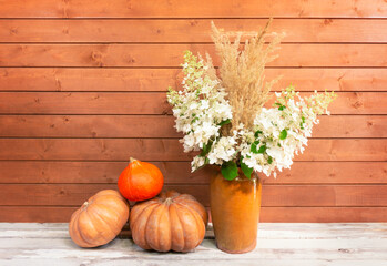 White hydrangea flowers and dry grass in clay vase and pumpkins on aged wooden table.