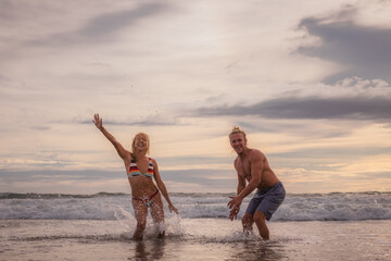 Happy Caucasian couple playing with water on the beach. Smiling and laughing young man and woman enjoying life. Sunset time. Cloudy sky. Ocean with waves. Vacation in Asia. Bali, Seminyak