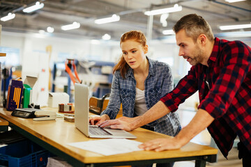 Young man and young woman using a laptop while working in a printing press office - obrazy, fototapety, plakaty