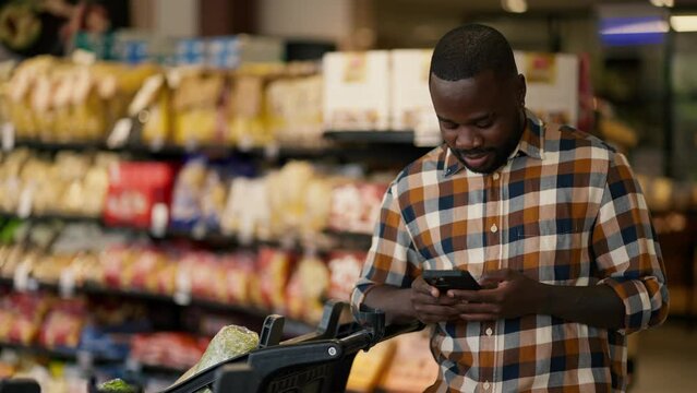 a man with Black skin color in a plaid shirt is typing and chatting on a black phone, next to him is a cart in a supermarket
